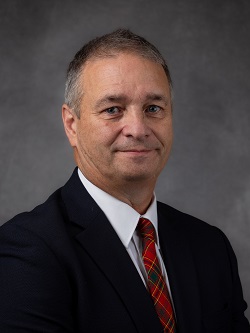 Councilor Mark Perterson wearing a dark blue suit with a red, green and yellow plaid tye, he has short dark brown hair. 