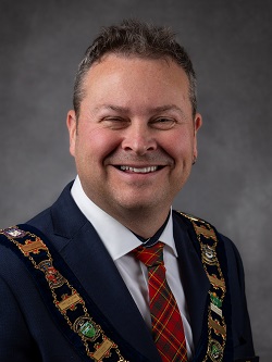 Warden Marcus Ryan wearing the Chain of office, a dark blue suit with a red, green and yellow plaid tie. 