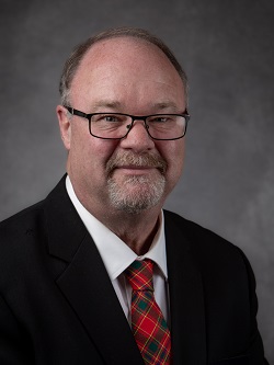Councilor Phil Schaefer, wearing a dark blue suit with a red, green and yellow plaid tye, he has short light brown hair. 